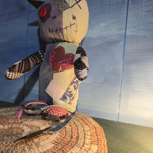 May include: A handmade fabric doll constructed from various patterned textile patches. The doll features a red button eye, a safety pin detail, and a prominent heart-shaped patch on its chest. It has two pointed ears and a multi-colored patchwork body.