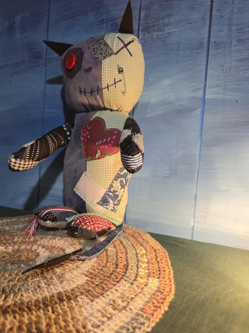 May include: A handmade fabric doll constructed from various patterned textile patches. The doll features a red button eye, a safety pin detail, and a prominent heart-shaped patch on its chest. It has two pointed ears and a multi-colored patchwork body.