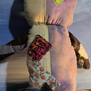 May include: A stuffed plush toy constructed from various fabric patches. The body features a light green gingham side and a purple tie-dye side. It includes a bright green rectangular patch, a floral print patch, and a dark patterned arm.