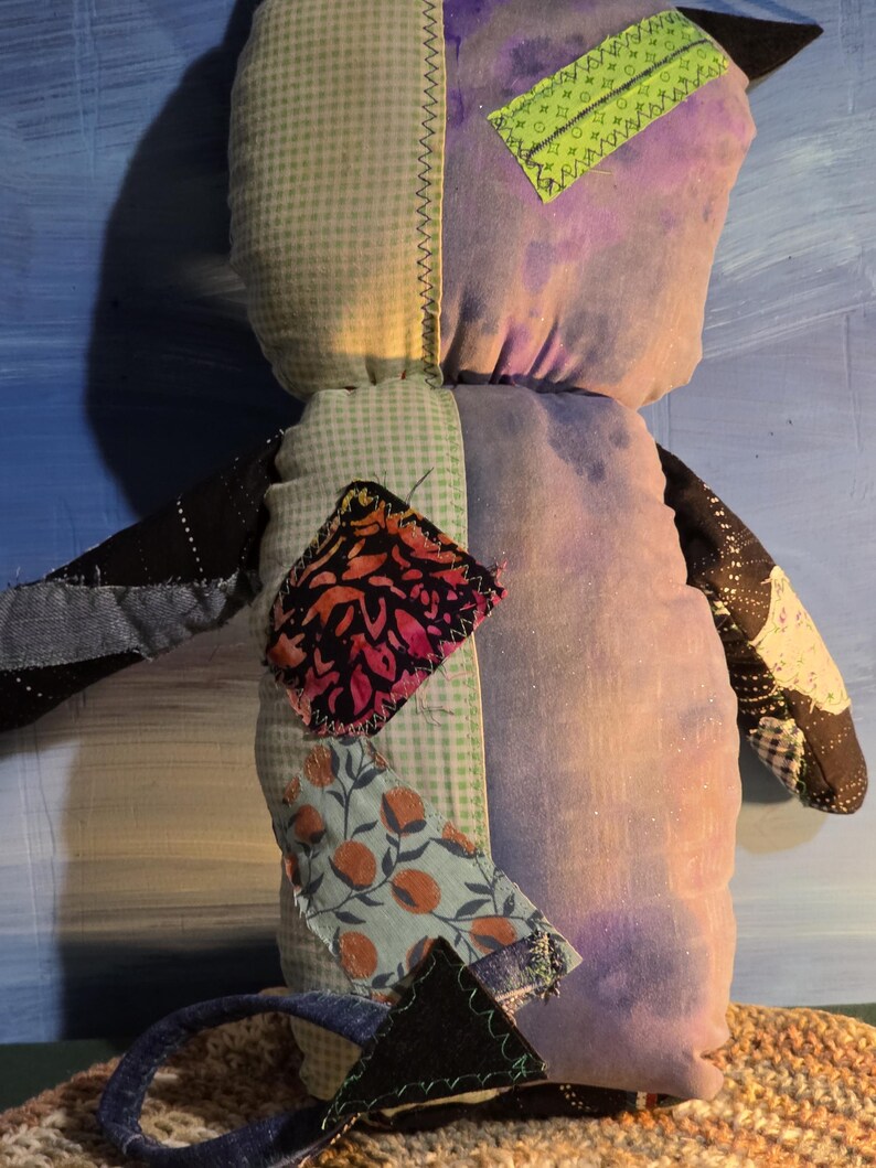 May include: A stuffed plush toy constructed from various fabric patches. The body features a light green gingham side and a purple tie-dye side. It includes a bright green rectangular patch, a floral print patch, and a dark patterned arm.