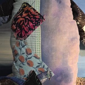 May include: A stuffed fabric toy featuring a patchwork design with a light green gingham section, a solid purple panel, and black sleeves. The toy is decorated with various fabric scraps, including a floral print and a dark patterned patch.