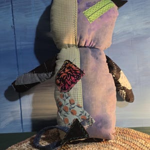 May include: A stuffed plush toy shaped like a cat with pointed ears and a tail. The body is constructed from various fabric patches including light green gingham, solid purple, black denim, and floral prints. A bright green rectangular patch is sewn on the head.