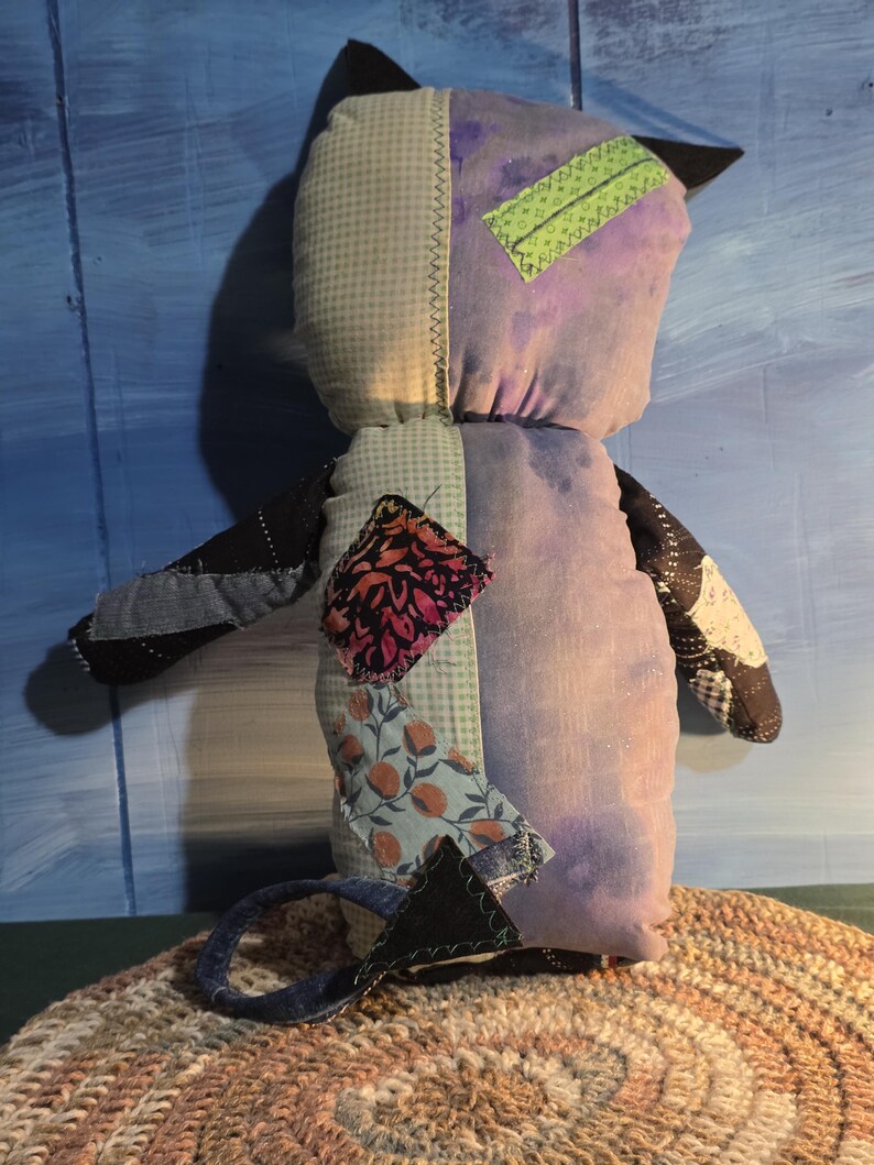 May include: A stuffed plush toy shaped like a cat with pointed ears and a tail. The body is constructed from various fabric patches including light green gingham, solid purple, black denim, and floral prints. A bright green rectangular patch is sewn on the head.