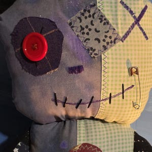 May include: A plush doll constructed from various fabric patches, including light blue, dark blue, and green gingham. It features a large red button eye, a stitched mouth, a safety pin, and a dark heart-shaped patch on the chest area.