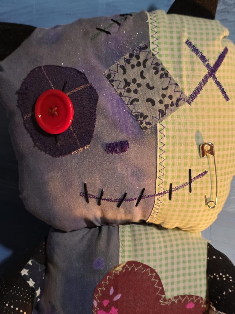 May include: A plush doll constructed from various fabric patches, including light blue, dark blue, and green gingham. It features a large red button eye, a stitched mouth, a safety pin, and a dark heart-shaped patch on the chest area.