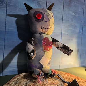 May include: A handmade plush doll constructed from various fabric patches in shades of blue, yellow, and black. The doll features a single red button eye, stitched mouth details, pointed ears, a heart-shaped chest patch, and a long pointed tail.