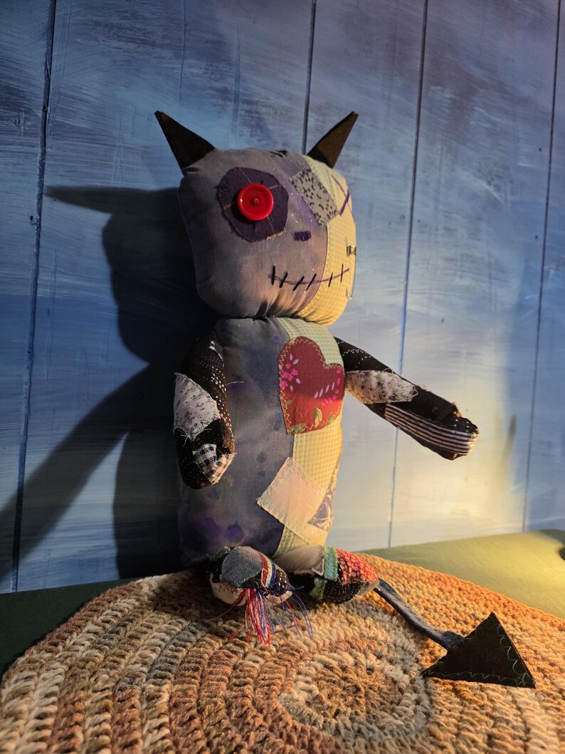 May include: A handmade plush doll constructed from various fabric patches in shades of blue, yellow, and black. The doll features a single red button eye, stitched mouth details, pointed ears, a heart-shaped chest patch, and a long pointed tail.