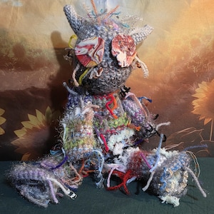 May include: A handmade crochet doll featuring a grey textured body with protruding ears and two large, round, multi-colored marbled buttons for eyes. The doll is adorned with various loose, colorful yarn strands and patches of different textures throughout.