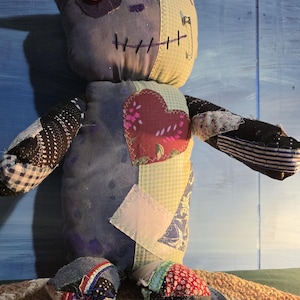 May include: A handmade plush doll constructed from various fabric patches in shades of blue, green, and red. The doll features a single red button eye, a stitched mouth, black pointed ears, and a prominent heart-shaped patch on the chest.