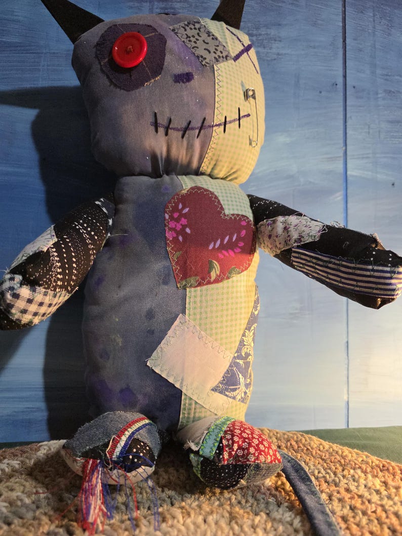 May include: A handmade plush doll constructed from various fabric patches in shades of blue, green, and red. The doll features a single red button eye, a stitched mouth, black pointed ears, and a prominent heart-shaped patch on the chest.