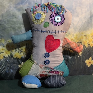 May include: A handmade rag doll with a gray body, featuring button eyes, a stitched mouth, and a red heart. The doll has patchwork details in various colors, including blue, green, and plaid, with colorful yarn hair and button accents. The doll is approximately 12 inches tall.