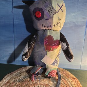 May include: A handmade plush doll featuring a patchwork design with various fabric patterns, a red button eye, a stitched mouth, and black pointed ears. The doll has a long tail with a black tip, a heart-shaped patch on the chest, and a safety pin.