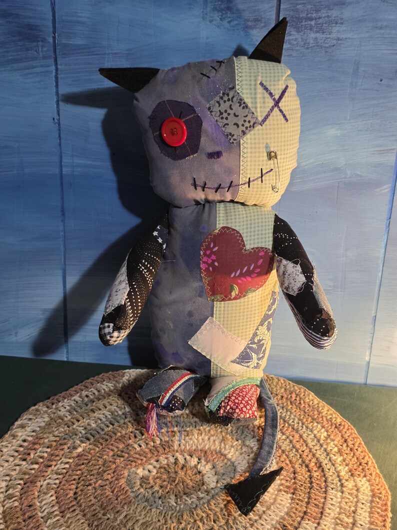 May include: A handmade plush doll featuring a patchwork design with various fabric patterns, a red button eye, a stitched mouth, and black pointed ears. The doll has a long tail with a black tip, a heart-shaped patch on the chest, and a safety pin.