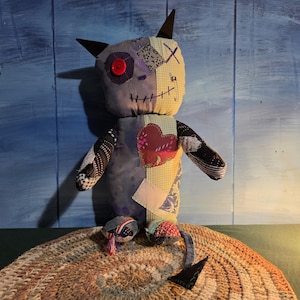 May include: A handmade fabric creature doll featuring a patchwork design with various patterns and colors, including a red button eye, a stitched mouth, a heart-shaped patch on the chest, pointed ears, and a long tail with a triangular tip.