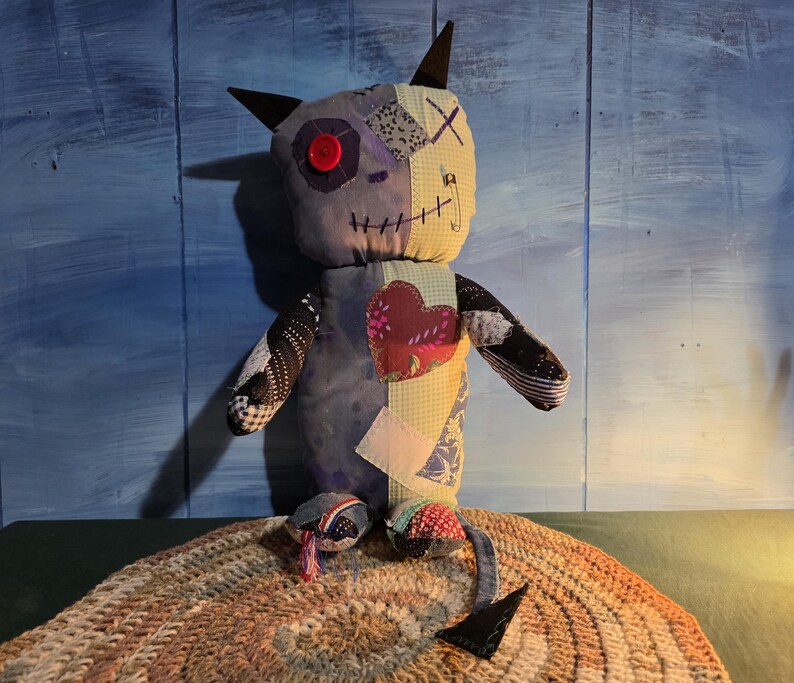 May include: A handmade fabric creature doll featuring a patchwork design with various patterns and colors, including a red button eye, a stitched mouth, a heart-shaped patch on the chest, pointed ears, and a long tail with a triangular tip.