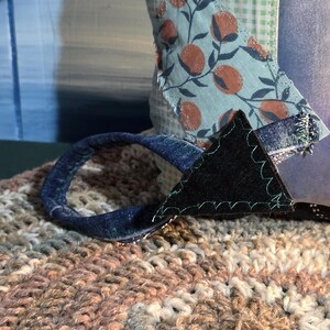 May include: A textile object featuring a light blue and white gingham fabric base, adorned with a patch of light blue fabric with a brown fruit print. A dark blue denim loop is attached with a black triangular fabric piece and green stitching.