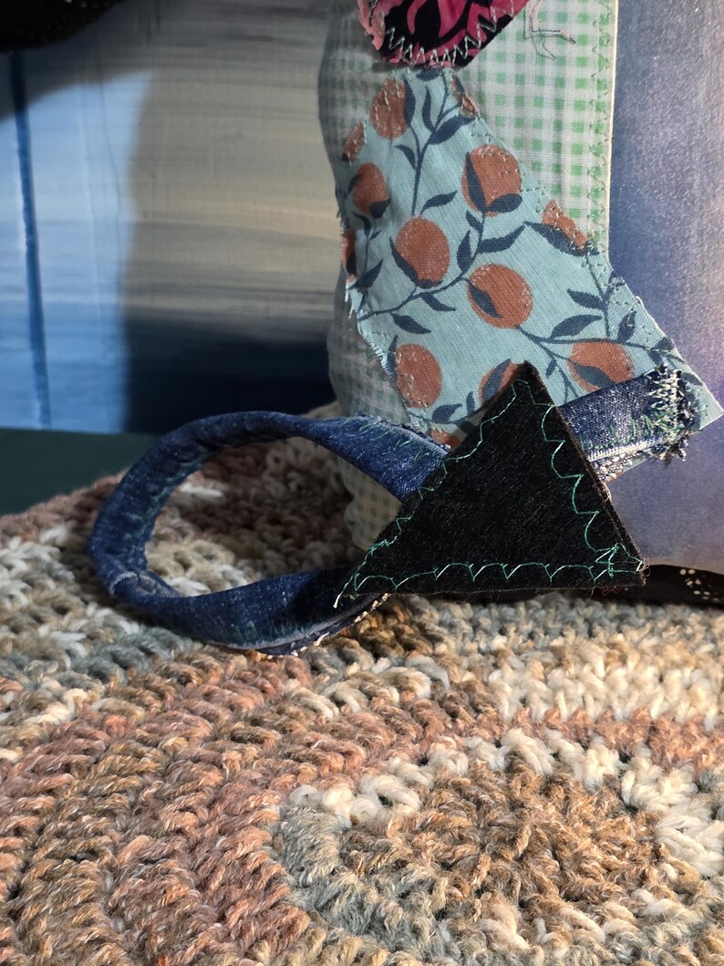 May include: A textile object featuring a light blue and white gingham fabric base, adorned with a patch of light blue fabric with a brown fruit print. A dark blue denim loop is attached with a black triangular fabric piece and green stitching.