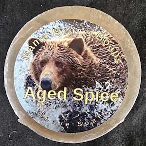 May include: Round shave soap with a label featuring a brown bear's face and the words "Mama Bear's Soaps" and "Aged Spice". The soap is a light beige color and appears to be wrapped in clear plastic.