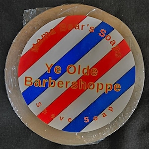 May include: Round shave soap with a red, white, and blue barber pole design. The soap is labeled "Mama Bear's Soaps" and "Ye Olde Barbershoppe shave soap."