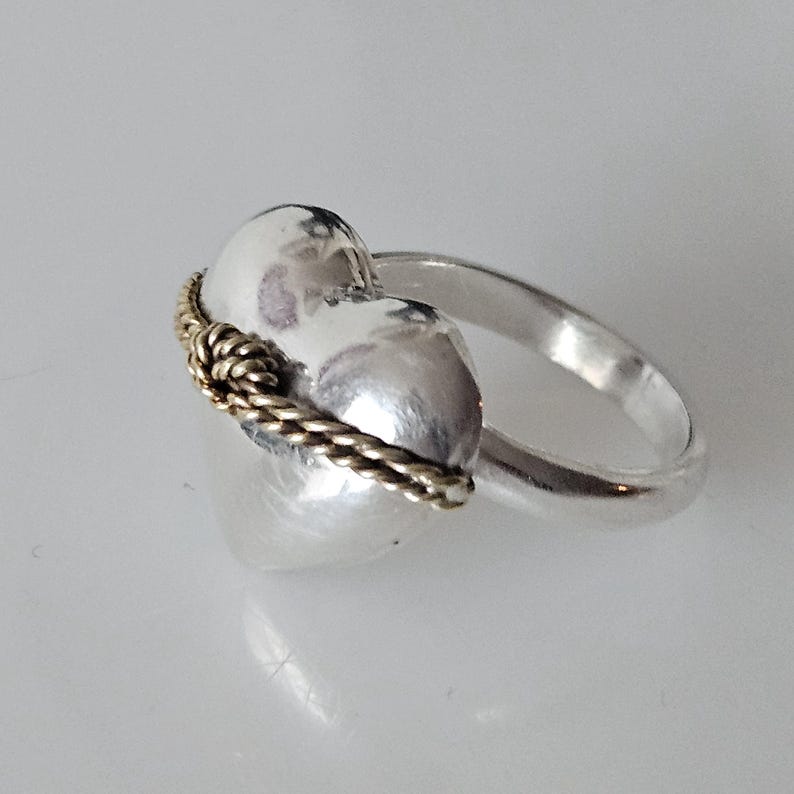 Op de afbeelding: Een zilveren ring in de vorm van een hart met een gouden touwontwerp rond de band.