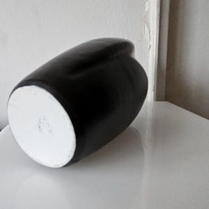 May include: A matte black ceramic vase with a smooth, rounded silhouette and a circular white base. The vessel features a curved, organic shape that tapers toward the top, showcasing a stark contrast between the dark exterior and the white bottom.
