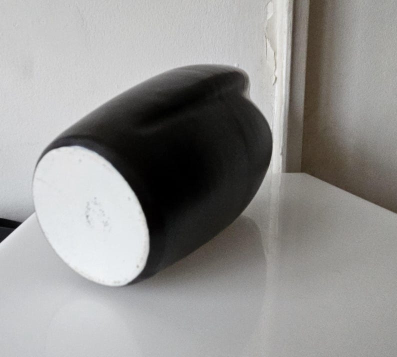 May include: A matte black ceramic vase with a smooth, rounded silhouette and a circular white base. The vessel features a curved, organic shape that tapers toward the top, showcasing a stark contrast between the dark exterior and the white bottom.