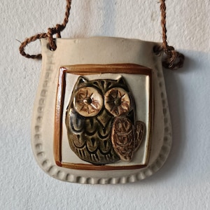 Könnte beinhalten: Eine cremefarbene, keramikartige Wandtasche in Eulenform mit braunen und grünen Details. Die Eule befindet sich in einem rechteckigen Rahmen mit strukturiertem Rand und einem braunen Seil zum Aufhängen. Die Eule hat große Augen und detaillierte Federn.