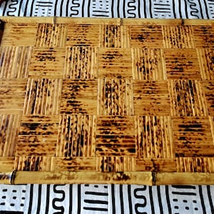 May include: Rectangular bamboo tray with a woven, checkered pattern. The tray has a natural, light brown color with darker, burnt-looking accents. The border is made of bamboo. The tray is on a black and white patterned fabric.
