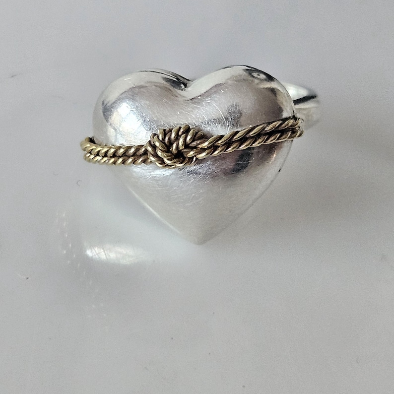Op de afbeelding: Een zilveren ring in de vorm van een hart met een gouden touwontwerp eromheen gewikkeld, waardoor een knoop bovenaan ontstaat.