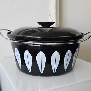 Può includere: Una pentola da cucina in smalto nero con coperchio e manici argentati. La pentola presenta un design a foglie bianche all'esterno. Il coperchio ha una piccola maniglia nera. Un oggetto vintage per la cucina.