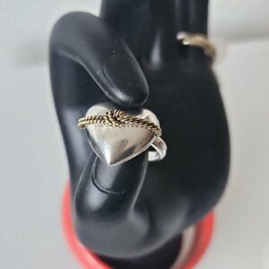 Op de afbeelding: Een zilveren ring in de vorm van een hart met een gouden touwontwerp rond het hart. De ring is op een zwart handmodel.