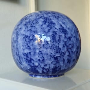 May include: A spherical ceramic vase featuring a deep blue base color with a mottled, cloud-like white glaze pattern across its surface. The object has a smooth, glossy finish and a rounded shape that tapers slightly toward a small circular base.