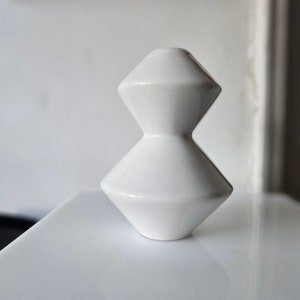 Puede incluir: Un jarrón de cerámica blanco con un diseño geométrico. El jarrón tiene una base ancha y un cuello estrecho.