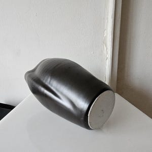 May include: A matte black ceramic vase with an organic, asymmetrical shape and a smooth, rounded body. The base of the vase is circular, flat, and unglazed, showing a light gray, textured clay surface that contrasts with the dark exterior finish.
