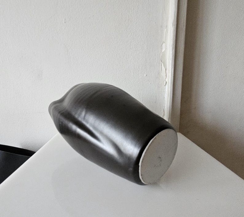 May include: A matte black ceramic vase with an organic, asymmetrical shape and a smooth, rounded body. The base of the vase is circular, flat, and unglazed, showing a light gray, textured clay surface that contrasts with the dark exterior finish.