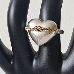 Op de afbeelding: Een zilveren ring in de vorm van een hart met een gouden touw eromheen gewikkeld.