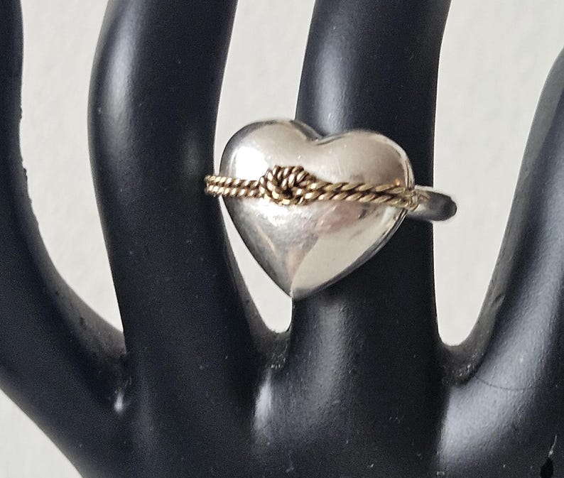 Op de afbeelding: Een zilveren ring in de vorm van een hart met een gouden touw eromheen gewikkeld.