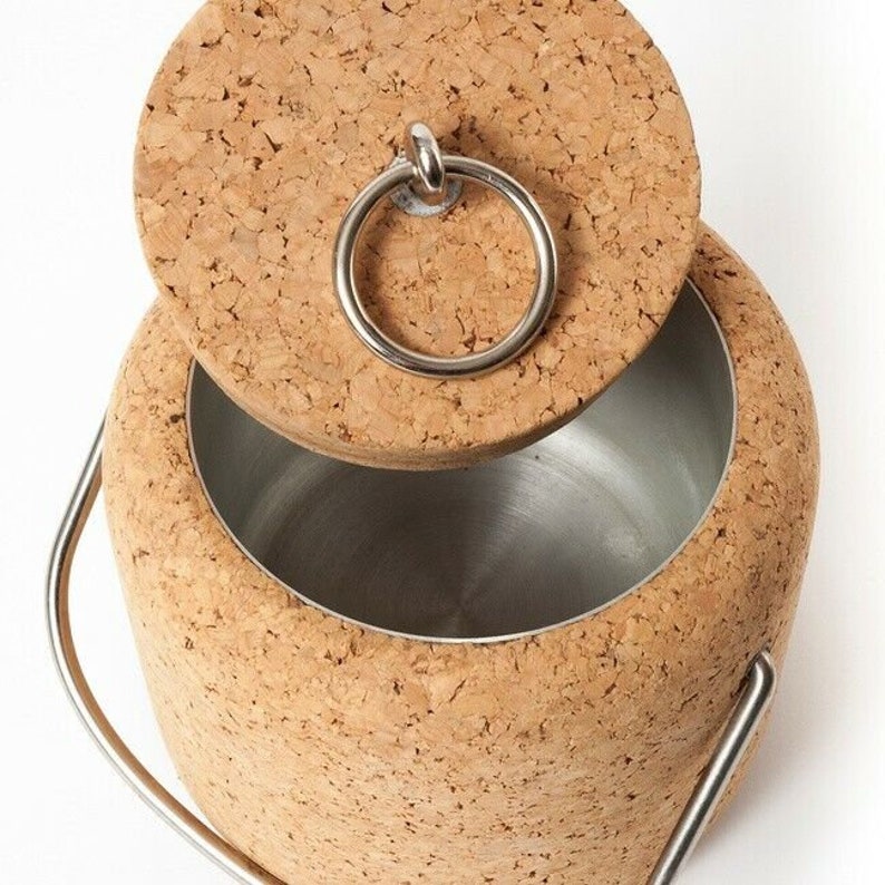 Puede incluir: Un cubo de hielo de corcho con un asa de metal y una tapa de corcho con un anillo de metal. El cubo est&aacute; vac&iacute;o y la tapa est&aacute; abierta.
