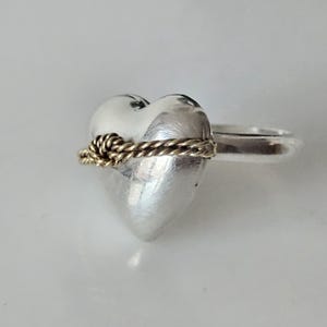 Op de afbeelding: Een zilveren ring in de vorm van een hart met een gouden touwontwerp rond het midden. De ring is een statement piece met een uniek en romantisch ontwerp.