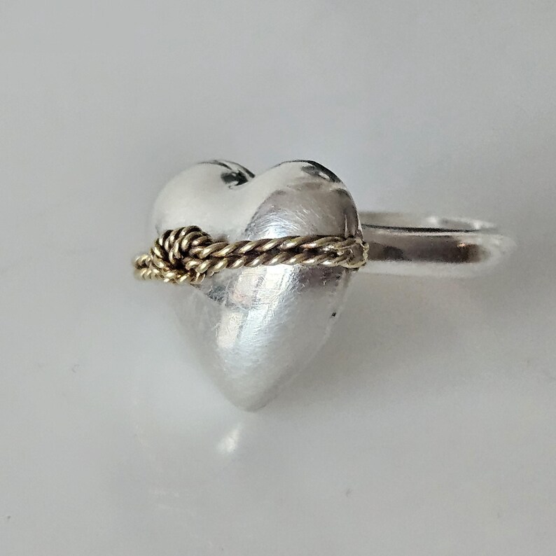 Op de afbeelding: Een zilveren ring in de vorm van een hart met een gouden touwontwerp rond het midden. De ring is een statement piece met een uniek en romantisch ontwerp.