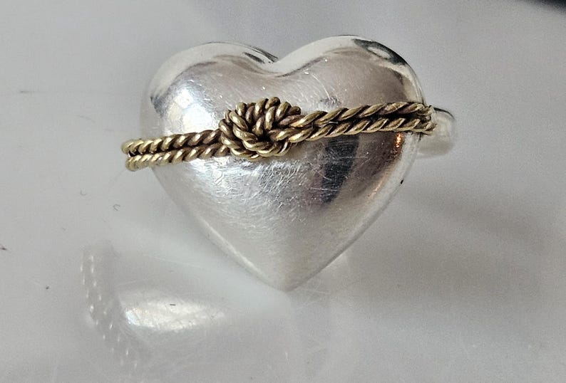 Op de afbeelding: Een zilveren ring in de vorm van een hart met een gouden touwontwerp eromheen gewikkeld, waardoor een knoop bovenaan ontstaat.