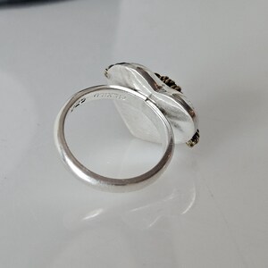 Op de afbeelding: Zilveren ring met een hartvormige steen gezet in een getextureerde zilveren bezel. De steen is omgeven door een dunne gouden draad. De ring is gemarkeerd met "925" op de binnenband.