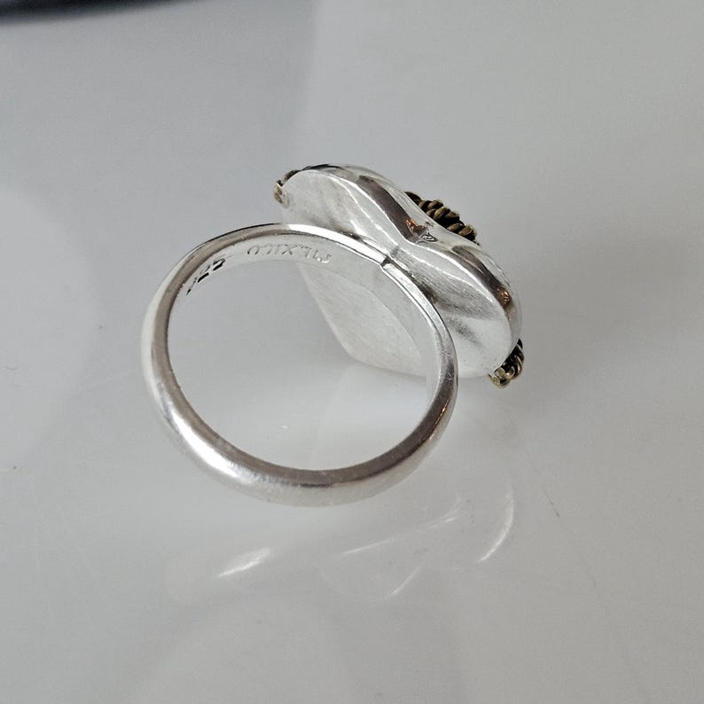 Op de afbeelding: Zilveren ring met een hartvormige steen gezet in een getextureerde zilveren bezel. De steen is omgeven door een dunne gouden draad. De ring is gemarkeerd met "925" op de binnenband.