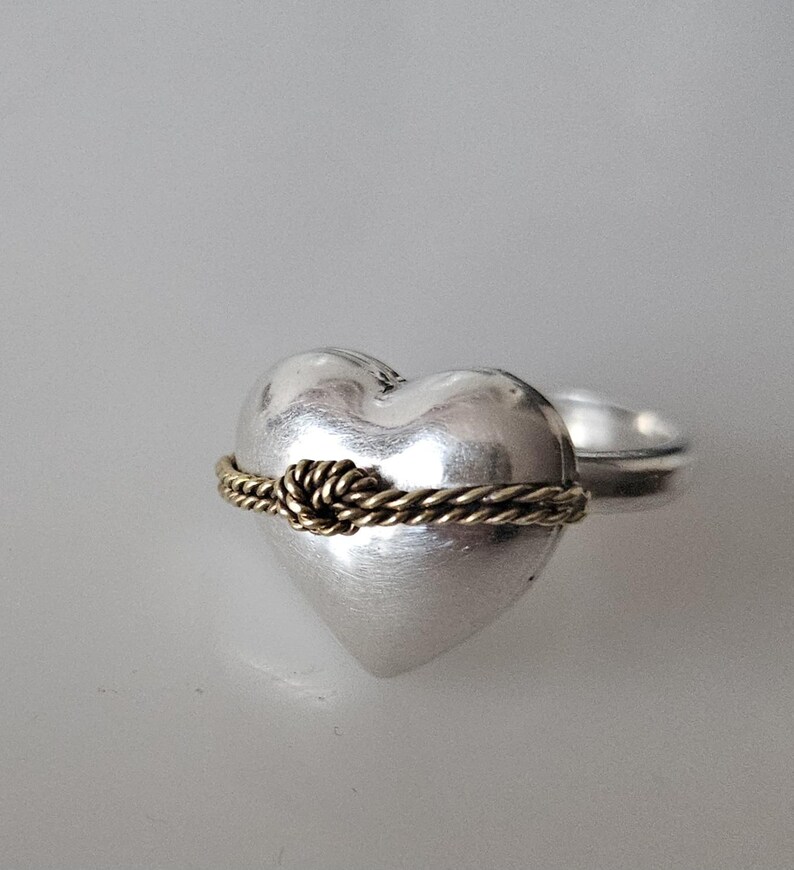 Op de afbeelding: Een zilveren ring in de vorm van een hart met een gouden touwontwerp rond het midden. Het touw is in een knoop geknoopt.