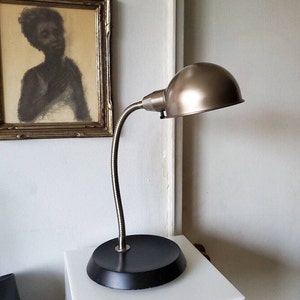 May include: A desk lamp with a metallic, dome-shaped shade and a flexible, silver-coloured neck. The lamp has a black, circular base. A framed portrait hangs in the background.
