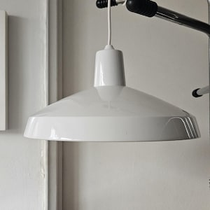 Può includere: Una lampada a sospensione bianca moderna con un paralume conico e una parte superiore cilindrica. La lampada è appesa a un cavo bianco fissato a un supporto metallico. La lampada è di colore bianco brillante e sembra essere in metallo.