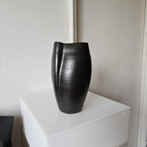 May include: A matte black ceramic vase with a smooth, rounded body and a distinct vertical indentation along one side. The vase features an asymmetrical, curved rim that dips slightly on the left, creating a fluid and organic silhouette.