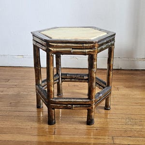 Puede incluir: Pequeña mesa auxiliar hexagonal con estructura de estilo bambú. El marco está pintado con un acabado dorado y bronce. La tapa de la mesa es de color beige claro. La mesa es una pieza decorativa.