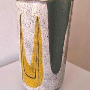 Könnte beinhalten: Eine hohe, gesprenkelte Keramikvase mit einem einzigartigen Design. Die Vase hat einen strukturierten, cremefarbenen Hintergrund mit schwarzen Sprenkeln. Kräftige gelbe Formen mit schwarzen Umrissen und ein dunkelgrüner Bereich sorgen für Kontrast.