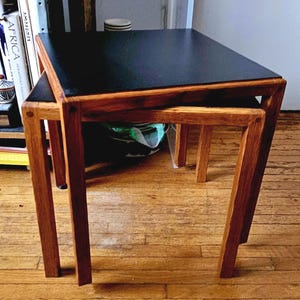 Puede incluir: Dos mesas nido de madera. La mesa superior tiene una superficie negra y la mesa inferior es visible debajo. Las mesas tienen un tono de madera cálido y están sobre un piso de madera noble. Las mesas son de forma cuadrada.